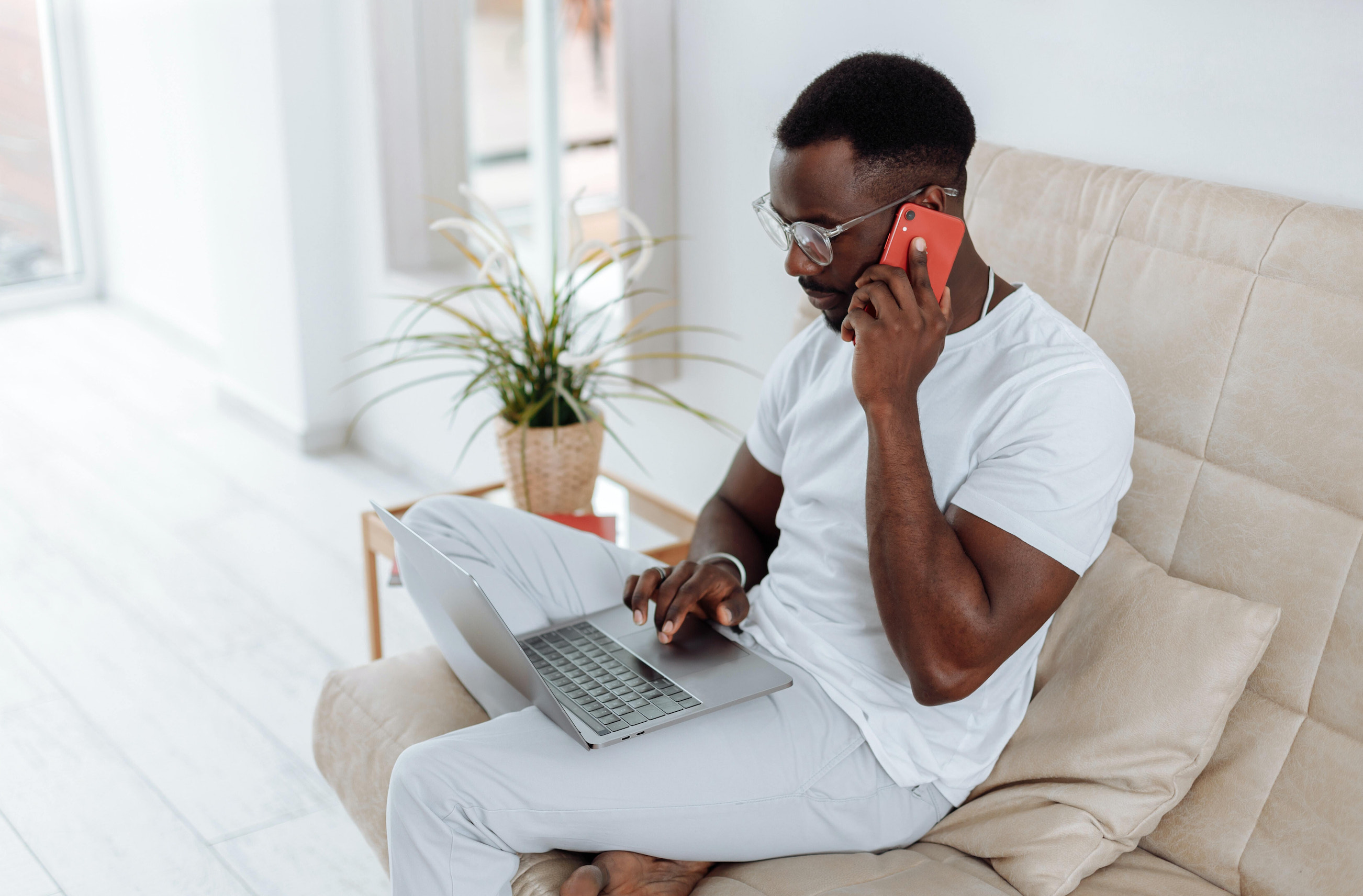 一名男子坐在沙发上,膝上放着一台笔记本电脑,把智能手机放在耳边