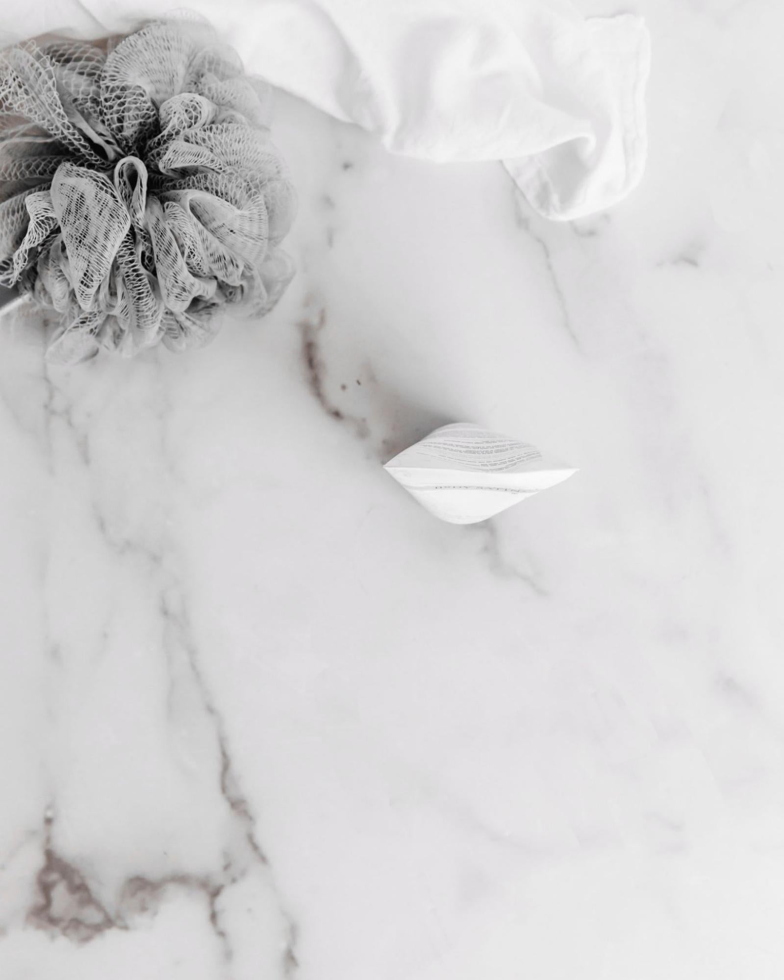 Same overhead shot of marble surface with luffa and towel but now with a bottle of hand cream in the center