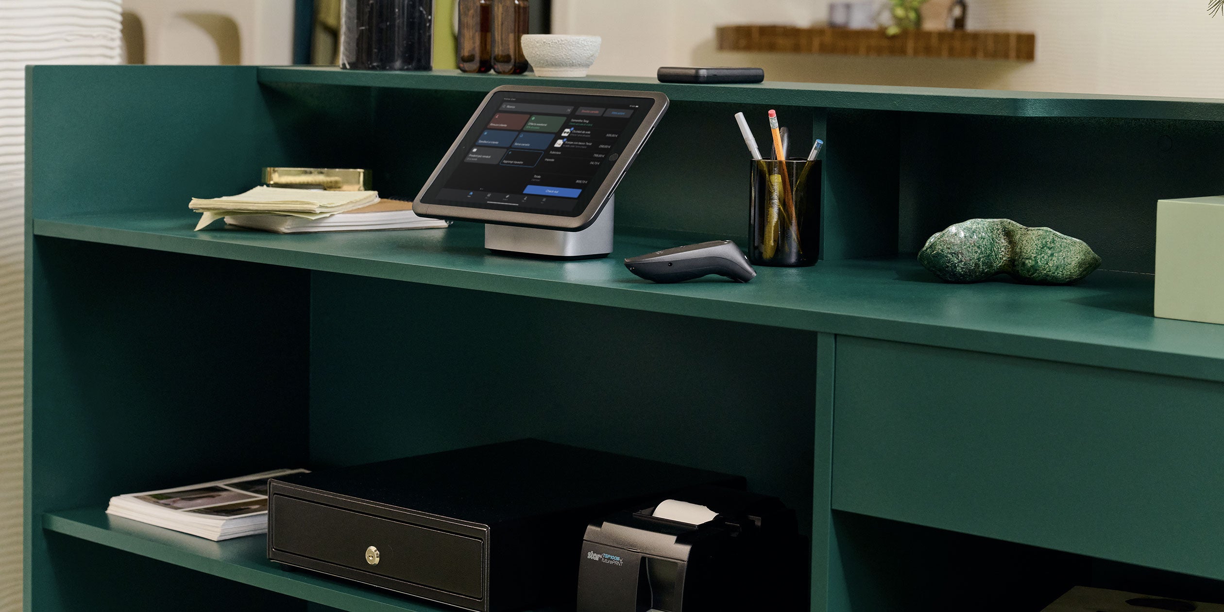 La cassa di un negozio vista da dietro, che mostra la configurazione di un POS con supporto per tablet, POS Go, cassetto portadenaro, stampante per ricevute e scanner per codici a barre.