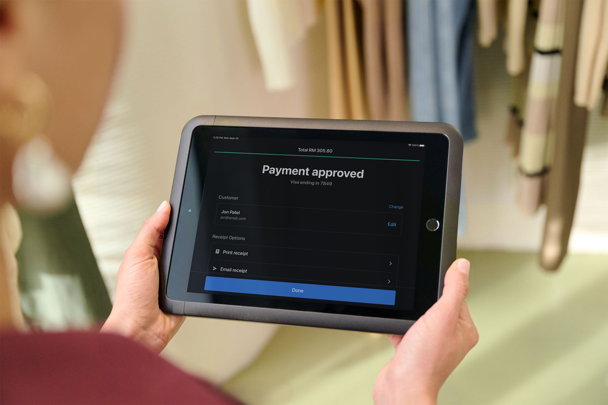 A sales associate holds a tablet running Shopify POS displaying the payment approved screen. On the screen: 